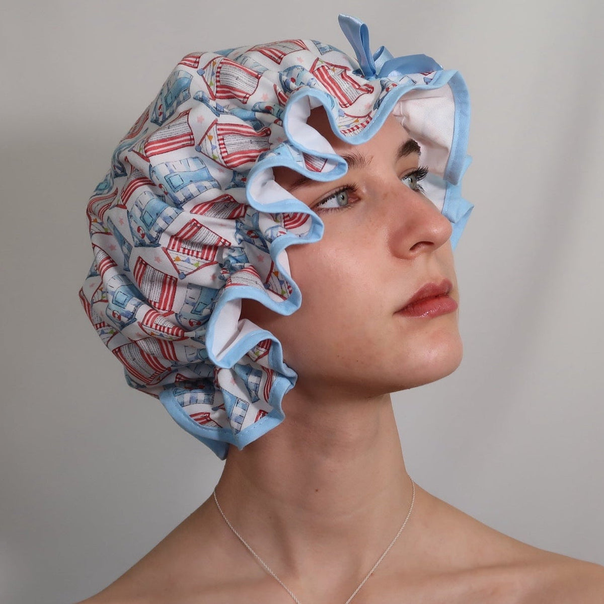 Woman wearing vintage style women's roomy cotton shower cap featuring multi-coloured beach huts on a white background. Trimmed sea blue with matching satin bow.