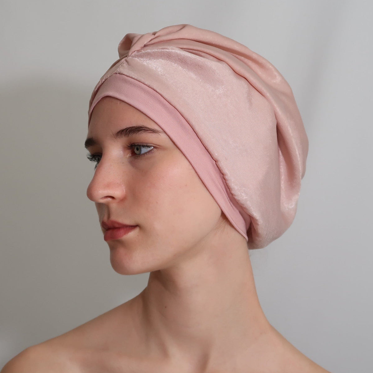 A woman in profile wearing a soft blush pink silk-lined sleep turban, photographed against a plain light gray studio background.