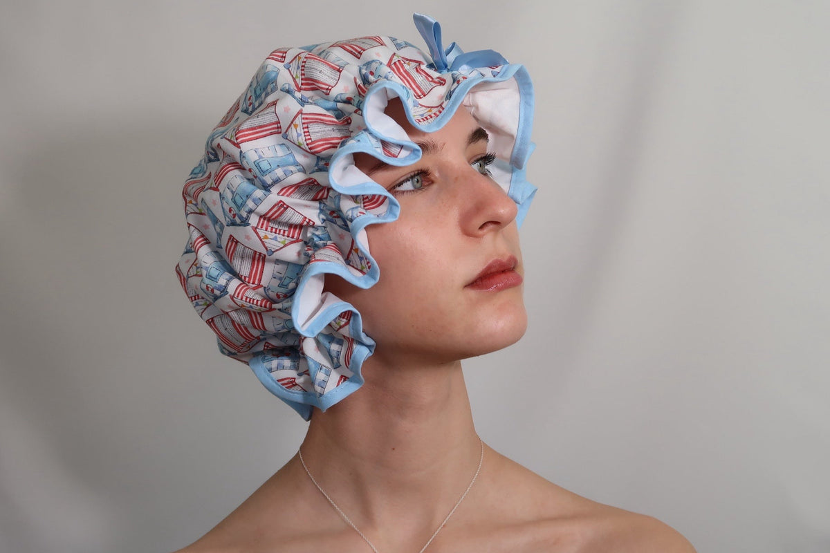 Woman wearing vintage style women's roomy cotton shower cap featuring multi-coloured beach huts on a white background. Trimmed sea blue with matching satin bow.