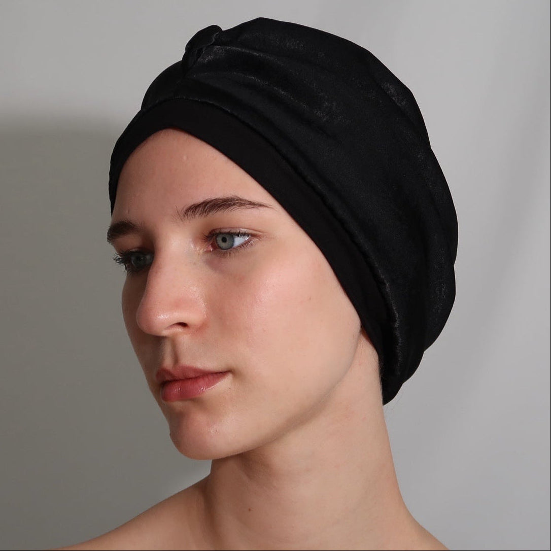 A woman wearing a black silk lined sleep turban, shown from the shoulders up against a plain light grey backdrop.