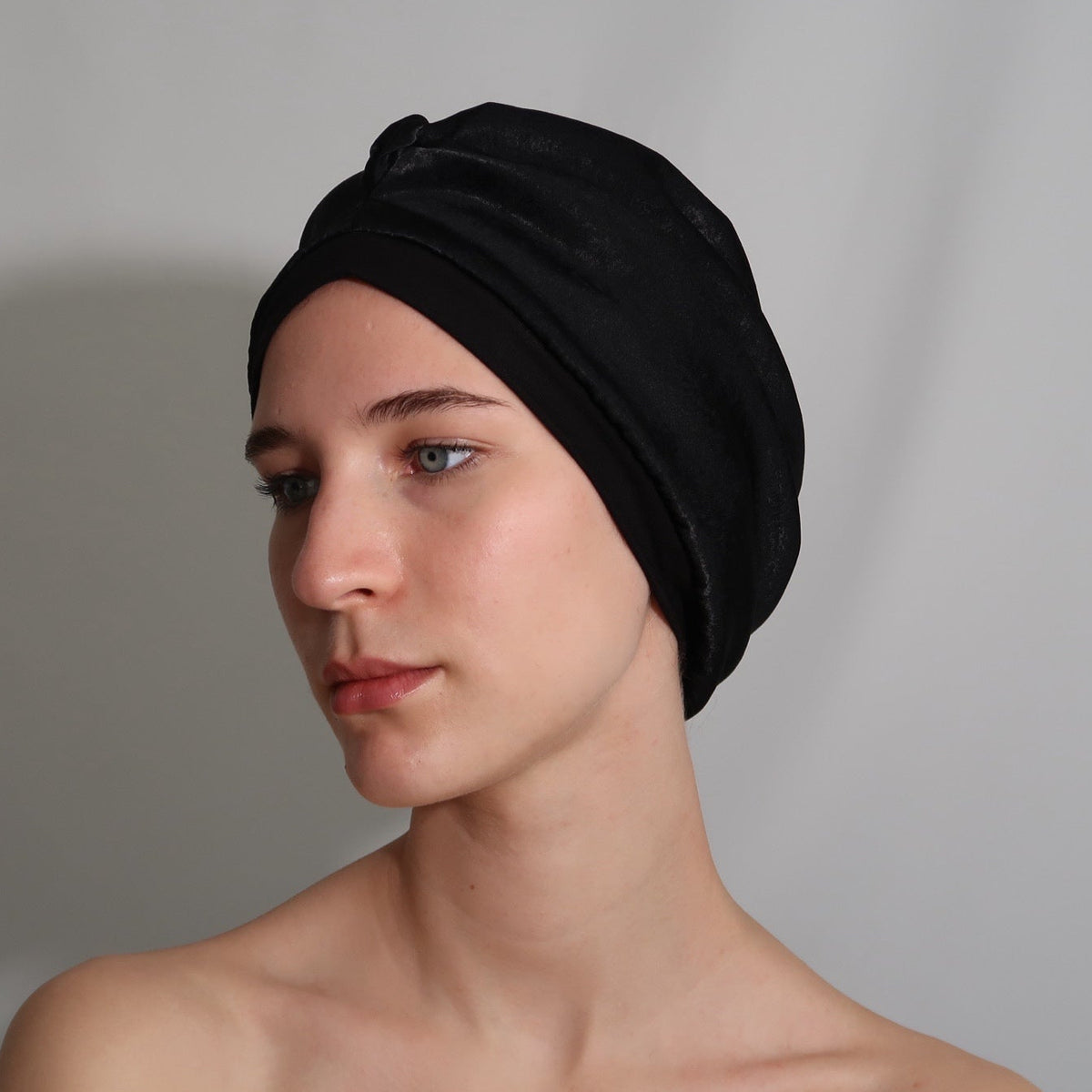 A woman wearing a black silk lined sleep turban, shown from the shoulders up against a plain light grey backdrop.
