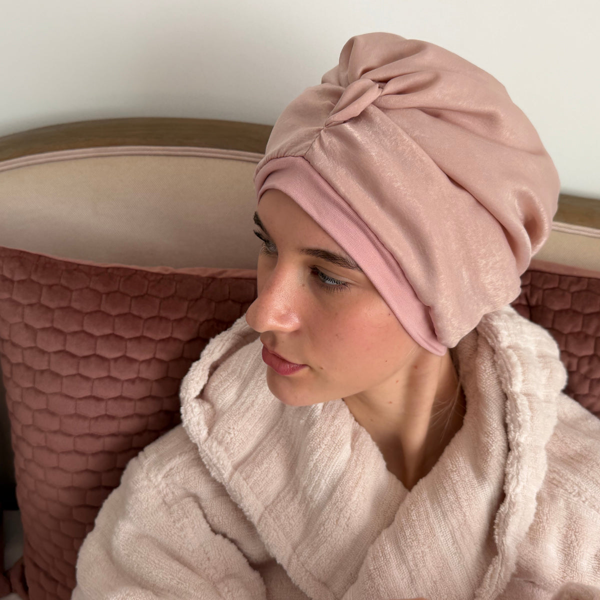 A woman wearing a dusky pink silk sleep turban with a matching light pink plush robe, sitting on a bed with pink pillows.