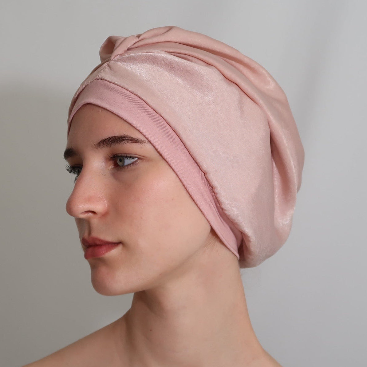 A woman in profile wearing a soft blush pink silk-lined sleep turban, photographed against a plain light gray studio background.