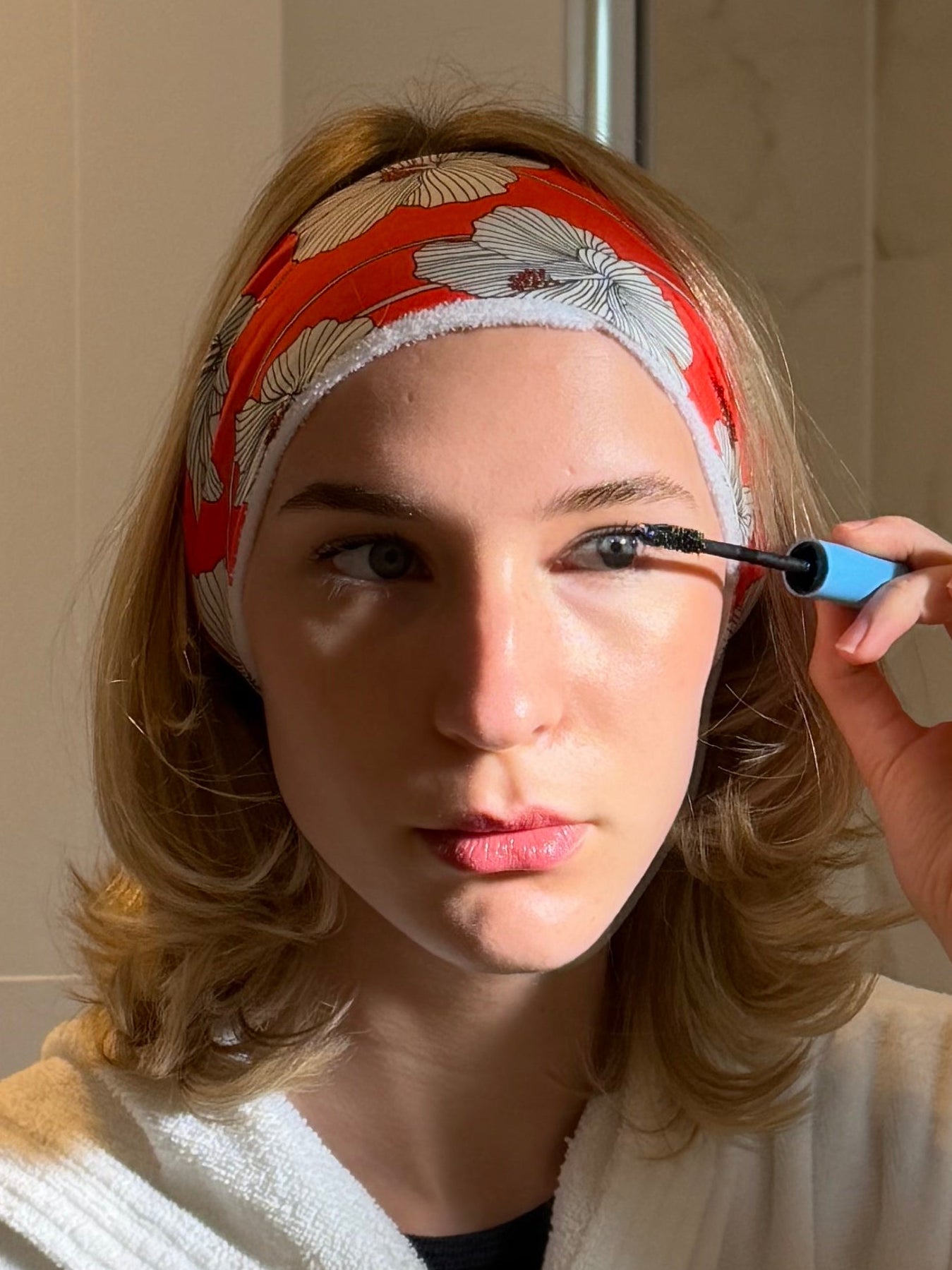 Woman applying makeup in front of a mirror in a bathroom wearing a red and white floral hairband