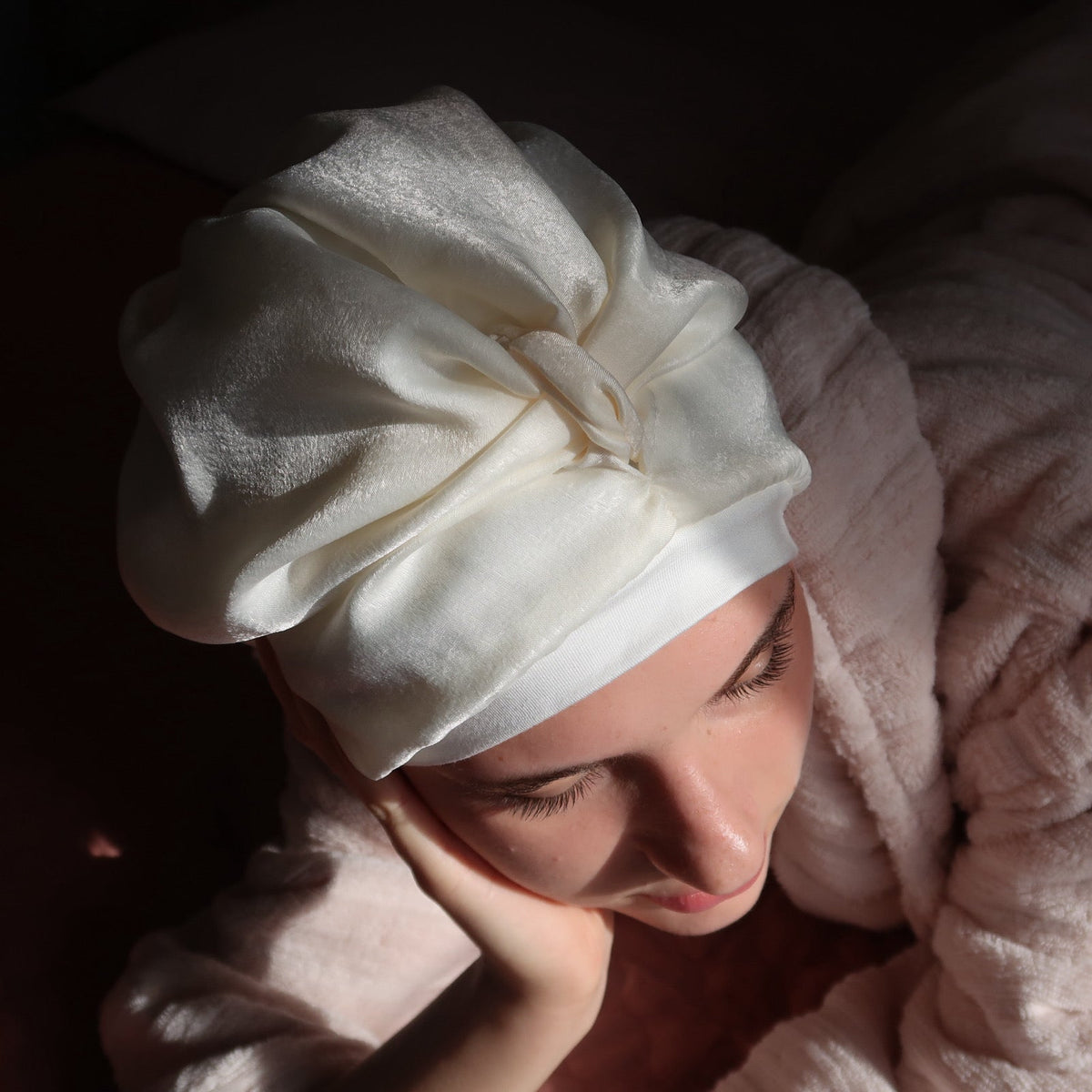 A woman lying down wrapped in a soft pink robe, wearing a cream-coloured silk sleep turban. The scene is lit with dramatic, warm sunlight, casting strong shadows against a dark background.