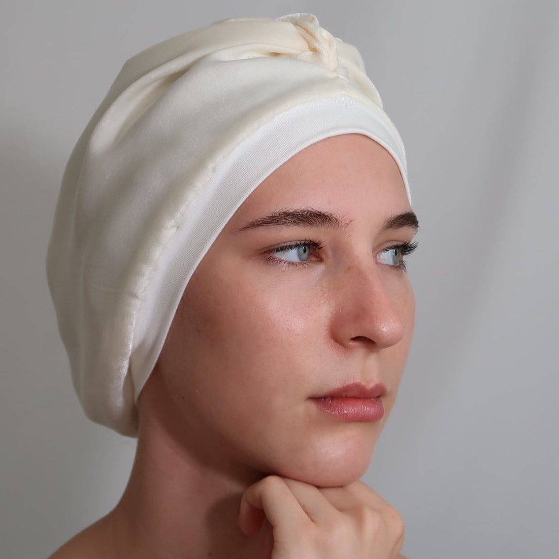A woman wearing a cream-coloured silk lined sleep turban, shown from the shoulders up against a neutral light grey background.