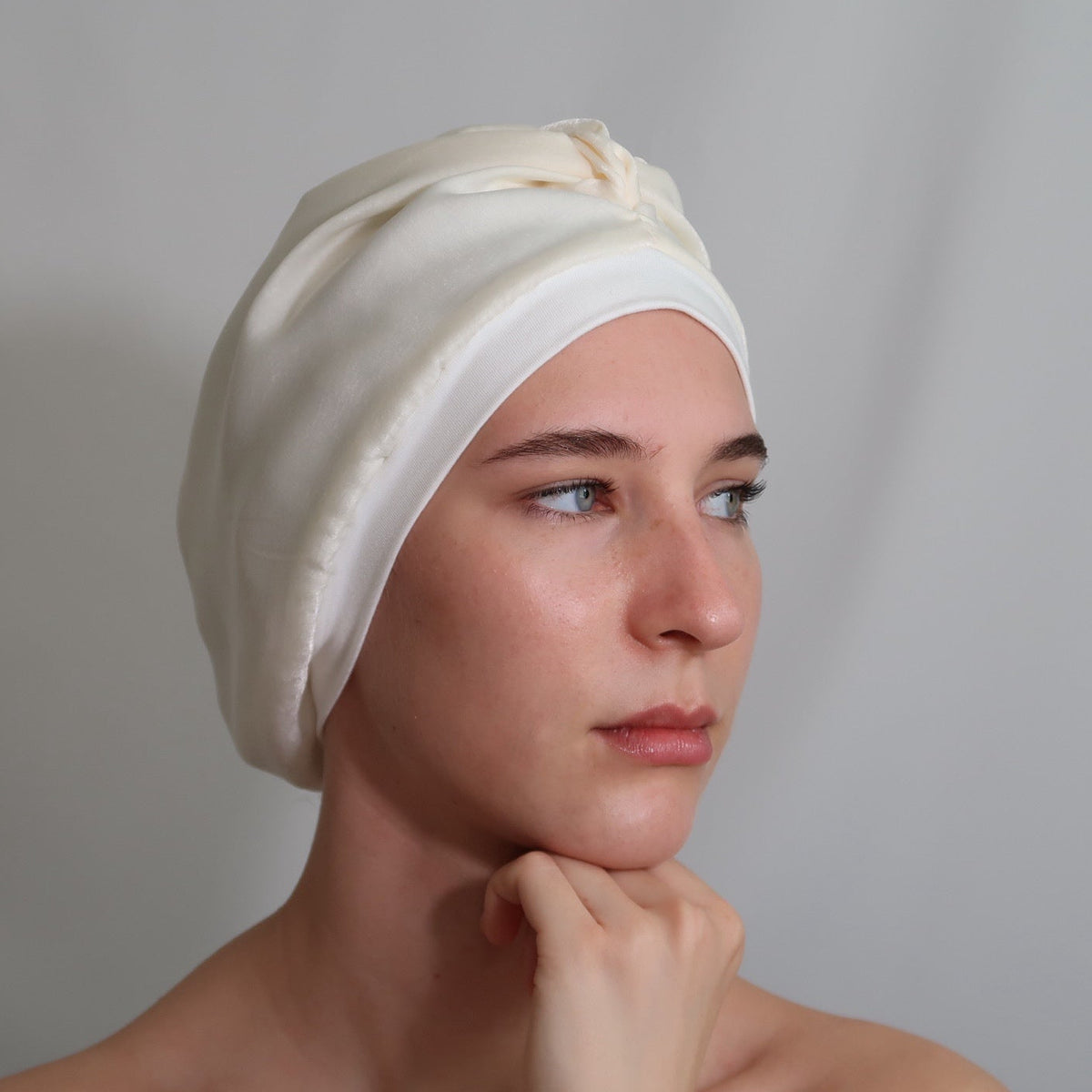 A woman wearing a cream-coloured silk lined sleep turban, shown from the shoulders up against a neutral light grey background.