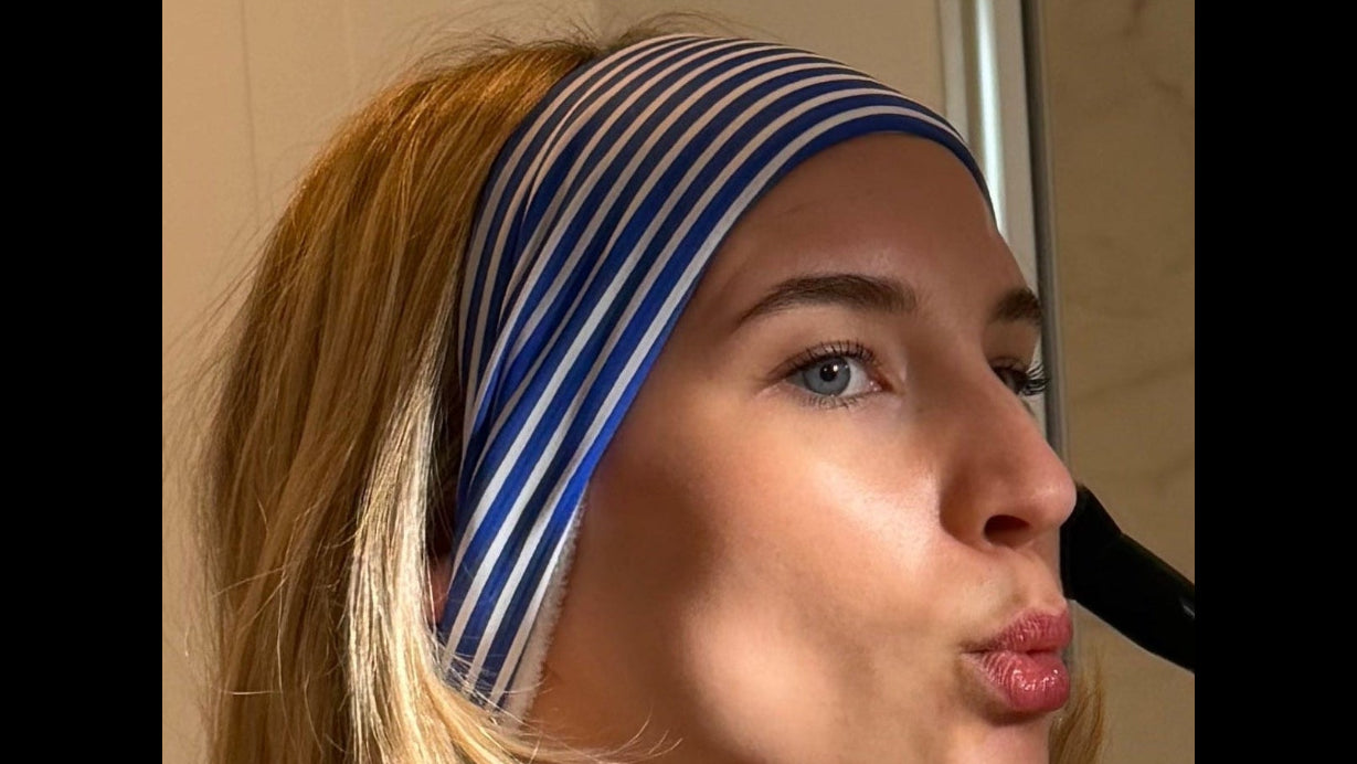 woman putting on make up in looking in bathroom mirror wearing a blue and white striped hairband