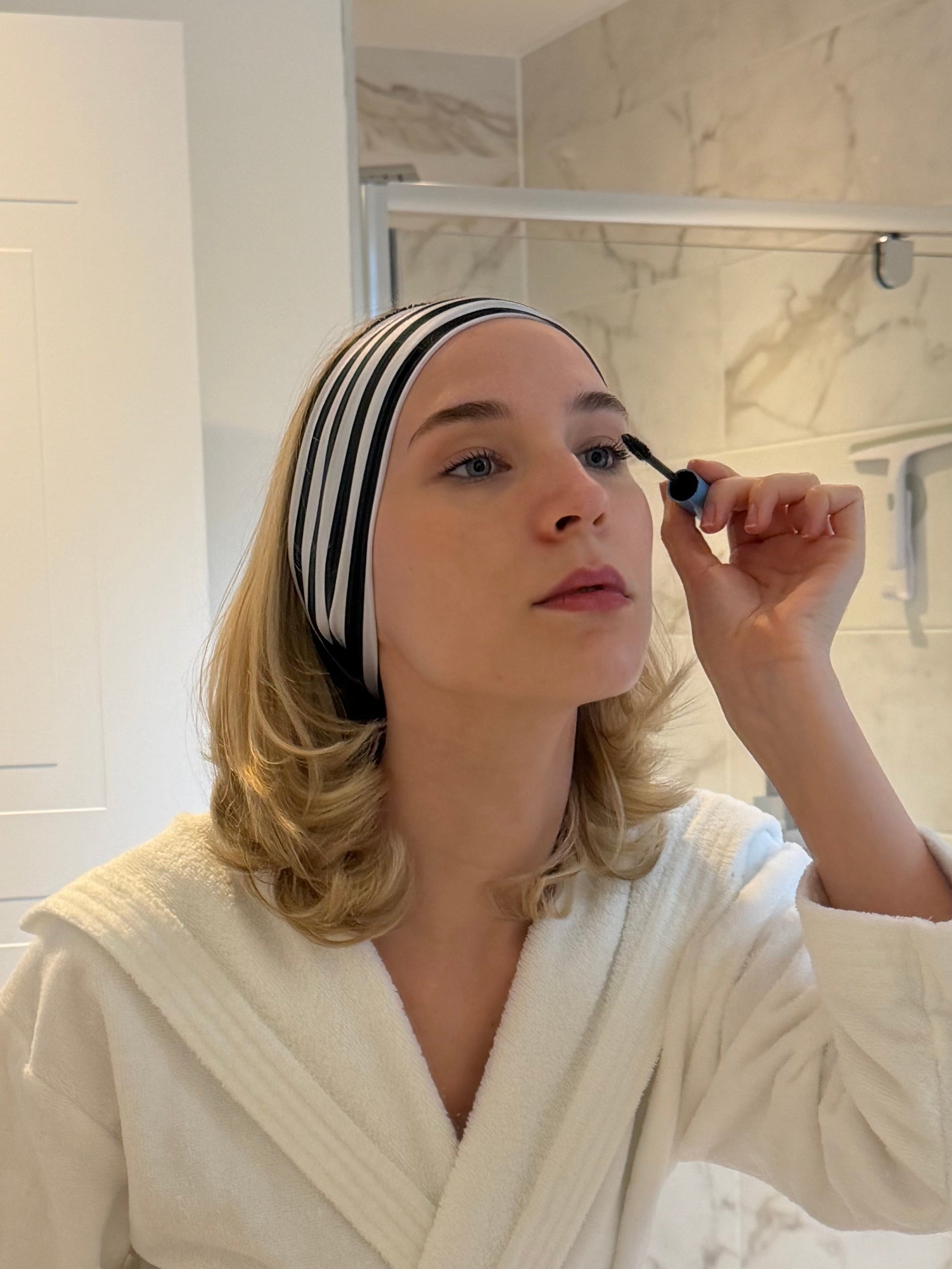 Woman applying mascara in front of a bathroom mirror wearing a robe and black and white striped hairband