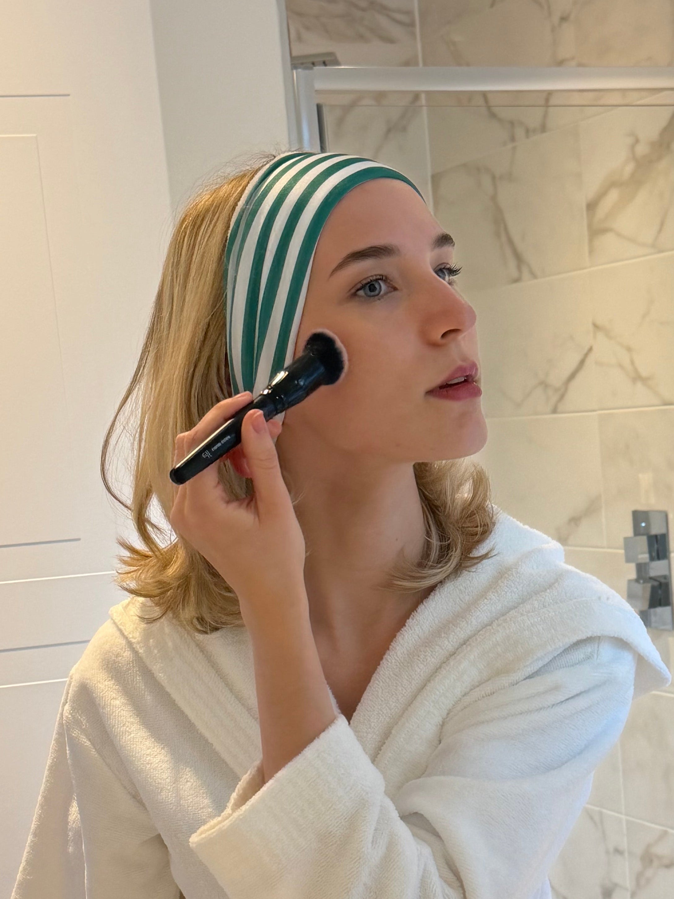Woman applying makeup in a bathroom mirror wearing a robe and green and white striped hairband