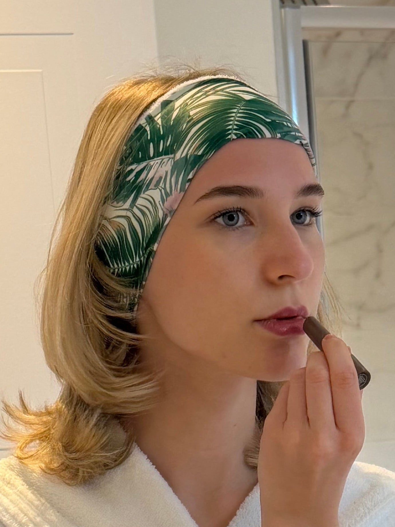 woman putting on lipstick in mirror wearing a green and white palm print hairband